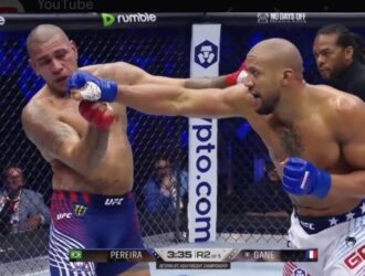 A high-resolution action shot from a UFC fight between Alex Pereira and Ciryl Gane, showing Gane landing a significant punch on Pereira's jaw while Pereira recoils, inside a packed arena with a visible broadcast graphic for Round 2.