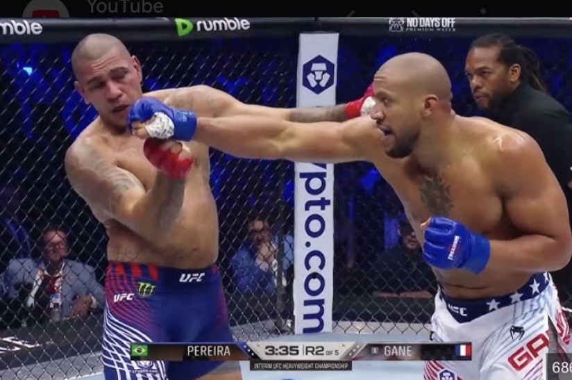 A high-resolution action shot from a UFC fight between Alex Pereira and Ciryl Gane, showing Gane landing a significant punch on Pereira's jaw while Pereira recoils, inside a packed arena with a visible broadcast graphic for Round 2.